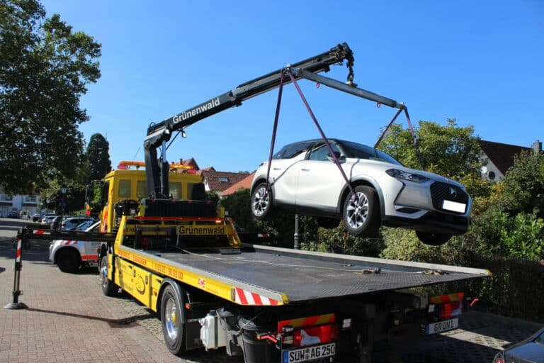 An einem sonnigen Tag wird in einem Wohnviertel ein weißes Auto von einem Kran auf einen gelben Abschleppwagen gehoben, was die Effizienz des Abschleppdienstes demonstriert.